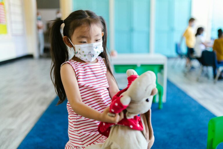 A young girl with pigtails wearing a face mask holding a stuffed toy in a classroom setting.