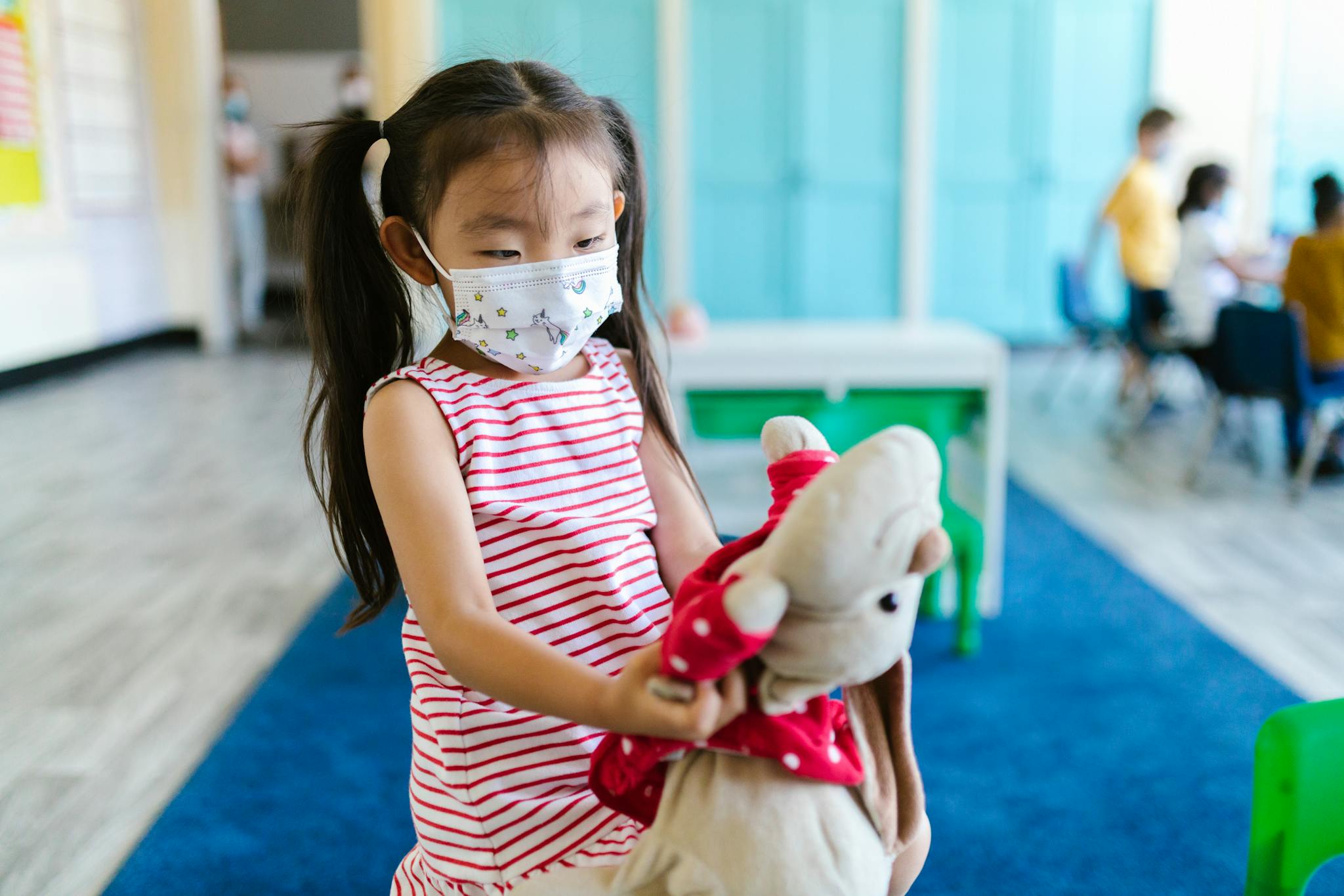 A young girl with pigtails wearing a face mask holding a stuffed toy in a classroom setting.